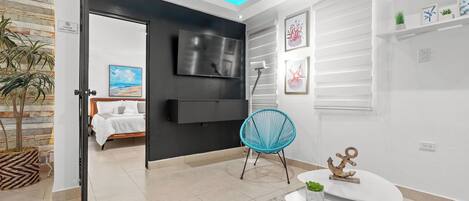 Una sala moderna con un televisor de pantalla plana, un sillón azul, una mesita con una planta y una cama al fondo.