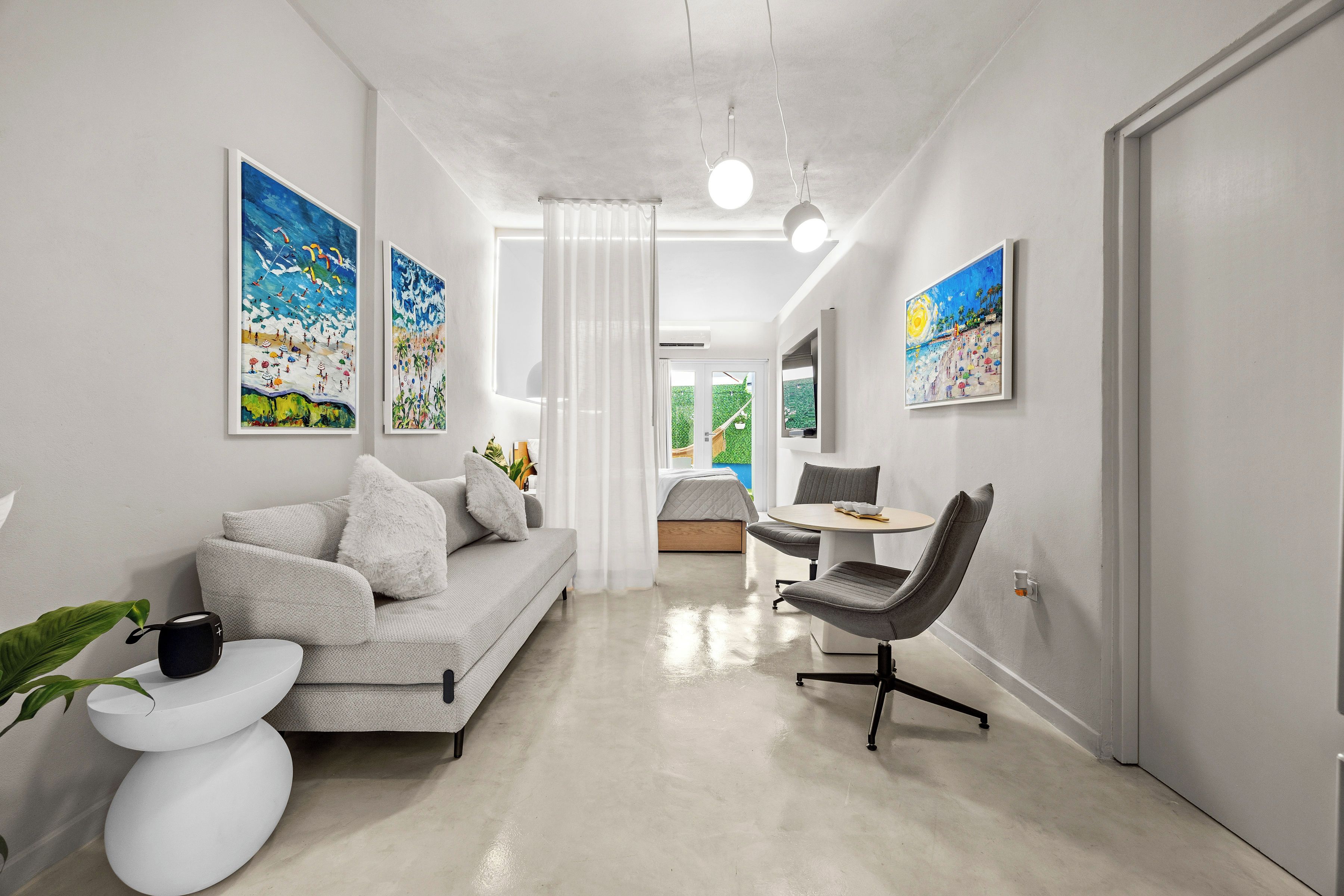 A modern living room with a sofa, a dining table, and a chair. There are paintings on the walls and a plant on the side table.