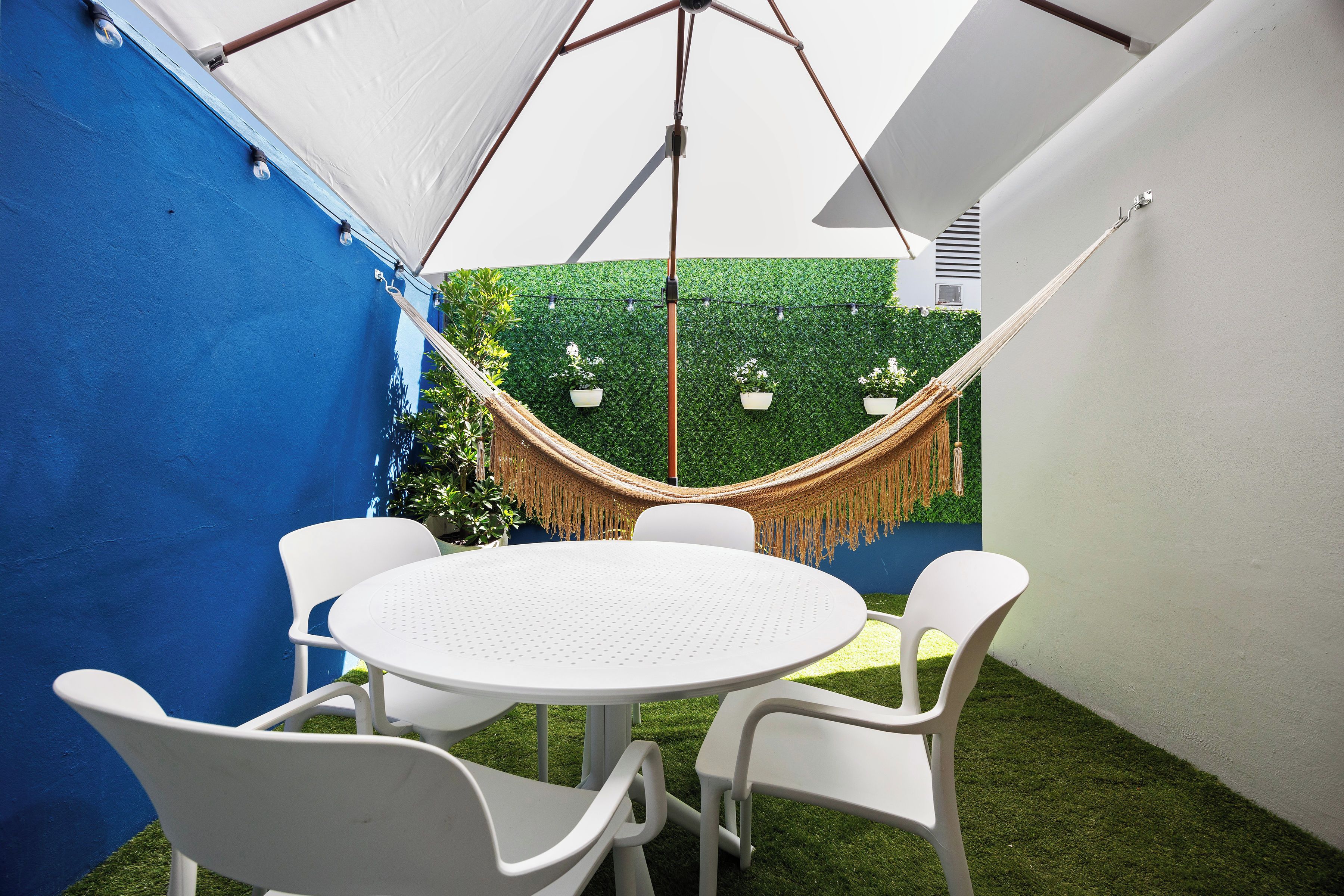A covered patio with a round table and white chairs, a hammock, and a green wall with hanging planters.