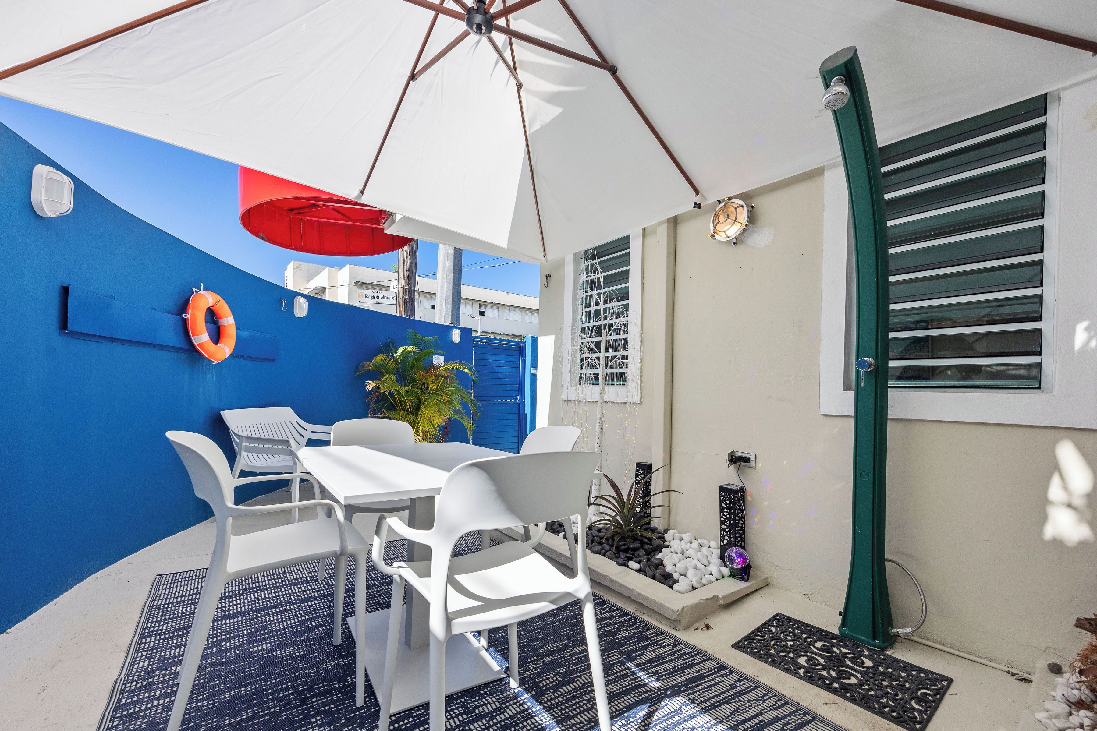 A covered outdoor seating area with white plastic chairs and a table, a life ring on the wall, and a blue and white wall.