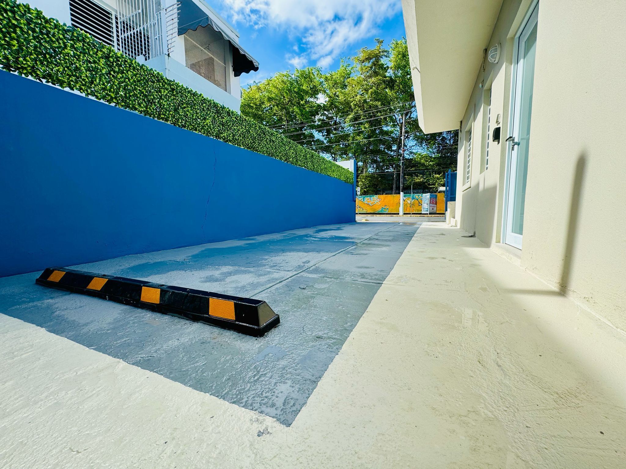 Un estrecho pasillo exterior con una pared azul en un lado y un edificio blanco en el otro. Una barrera negra y amarilla está colocada en el suelo.