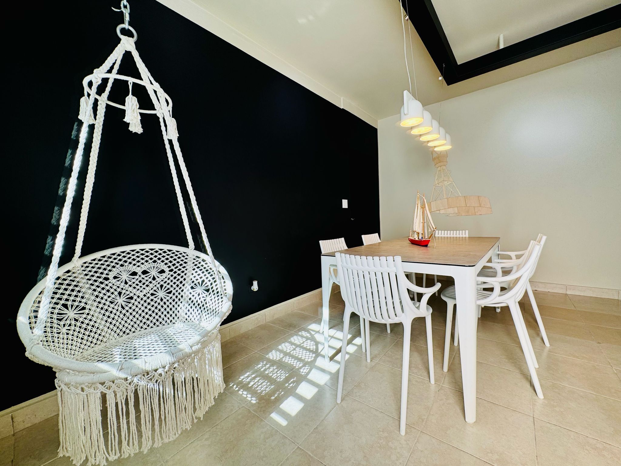 A dining area with a white macramé chair, white chairs, a wooden table, and a hanging light fixture.