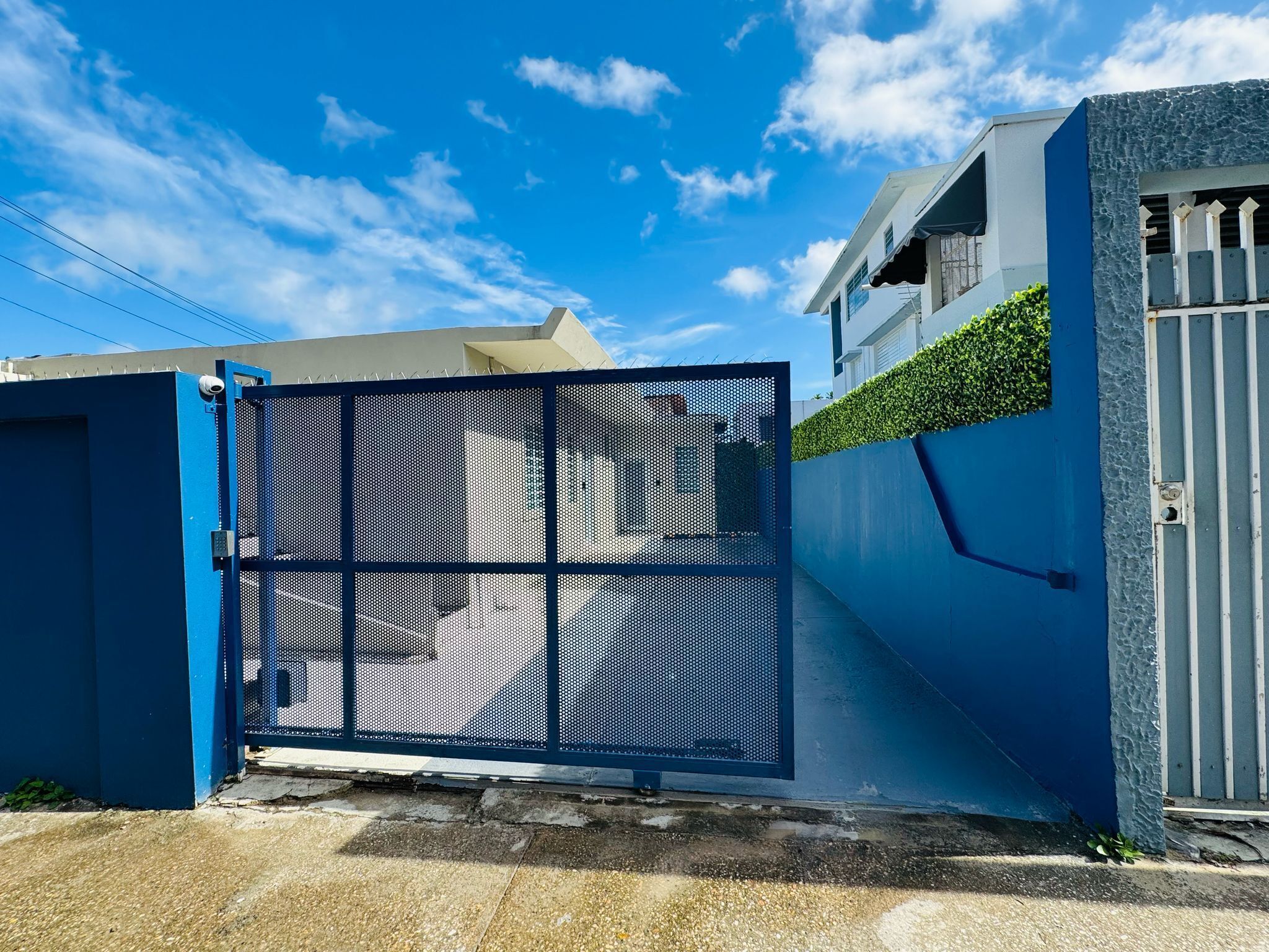 A gated entrance with a blue metal gate, a concrete pathway, and a building with a white facade and a green hedge.