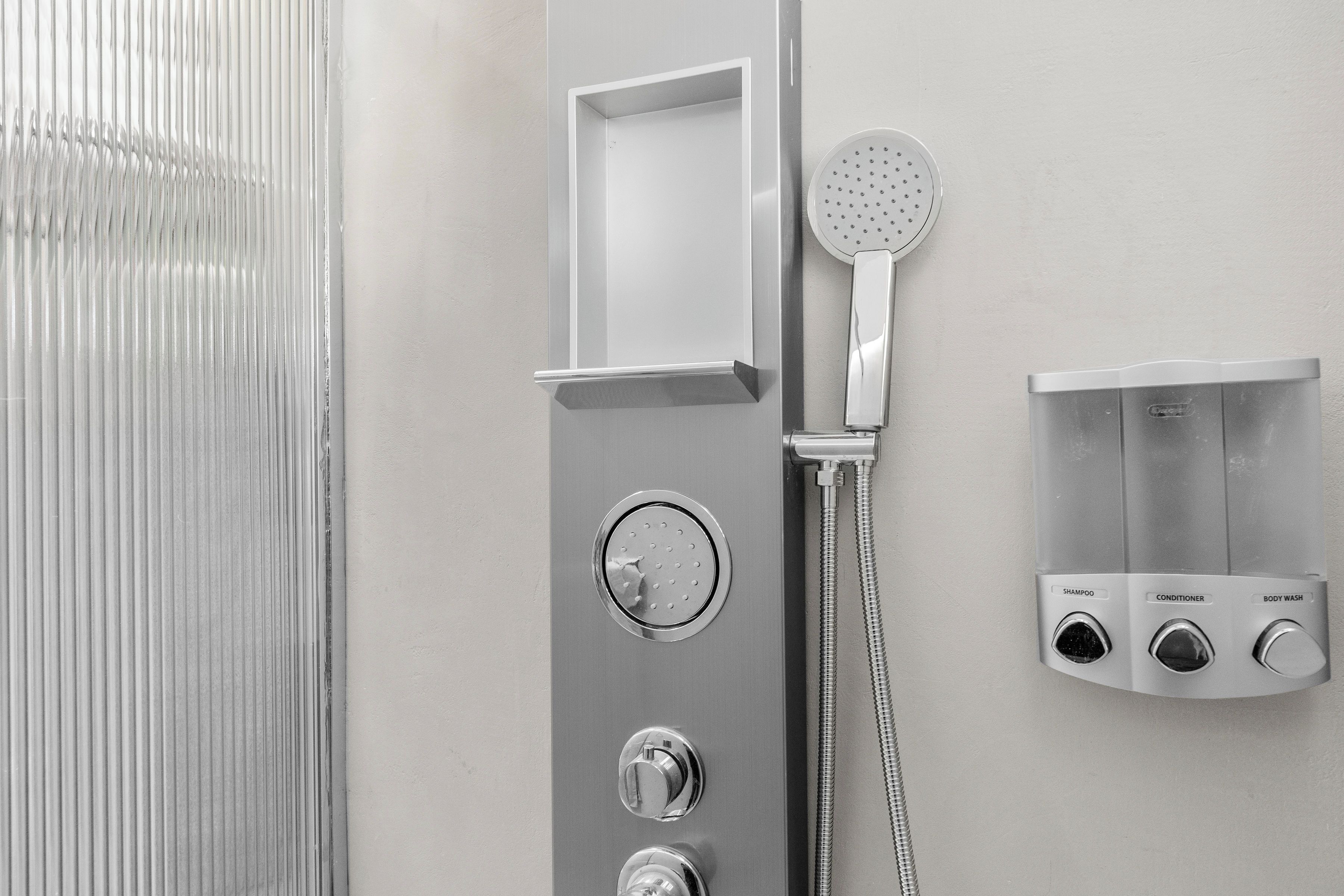 A modern shower with a handheld showerhead, a wall-mounted control panel with buttons for shampoo, conditioner, and body wash, and a small shelf above the control panel.