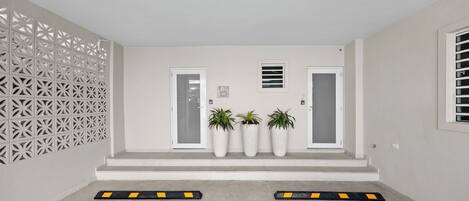 Una entrada limpia y moderna, con puerta blanca, plantas en macetas y un panel decorativo en la pared.