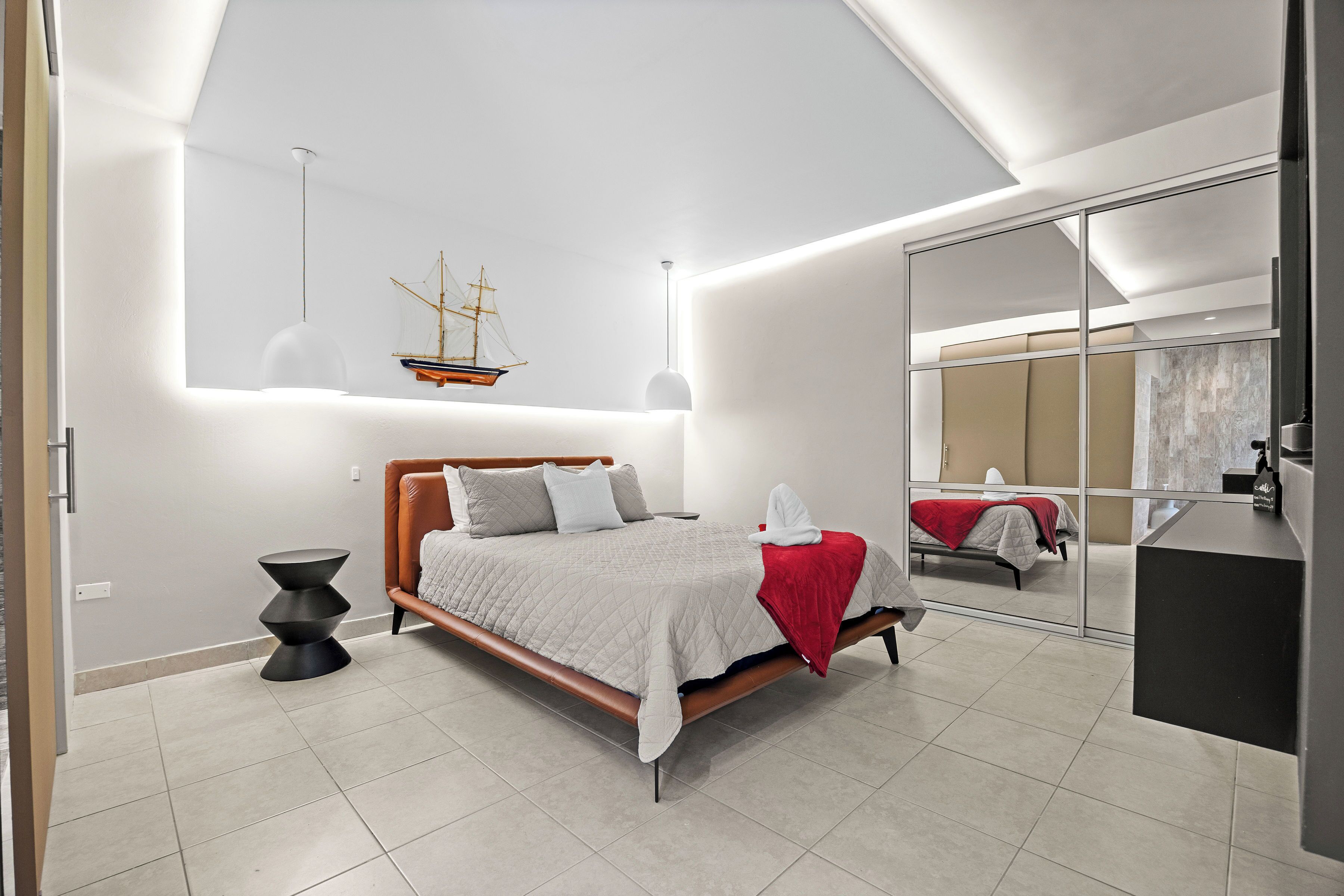A modern bedroom with a bed, a side table, a hanging ship model, and a large mirror.