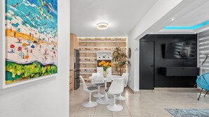 A modern dining area with a round table and white chairs, a large abstract painting on the wall, and a flat-screen TV mounted on the wall. - THE BUOY (San Juan)