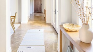 Un pasillo amplio con una puerta de madera al final, una alfombra blanca y un florero con plantas secas sobre una consola de madera.