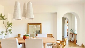 Un comedor con una mesa de madera, sillas blancas y un espejo grande encima. Hay lámparas colgantes y una planta sobre la mesa.