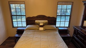 A bedroom with a large bed, two bedside tables, a dresser, and two windows with a view of trees.