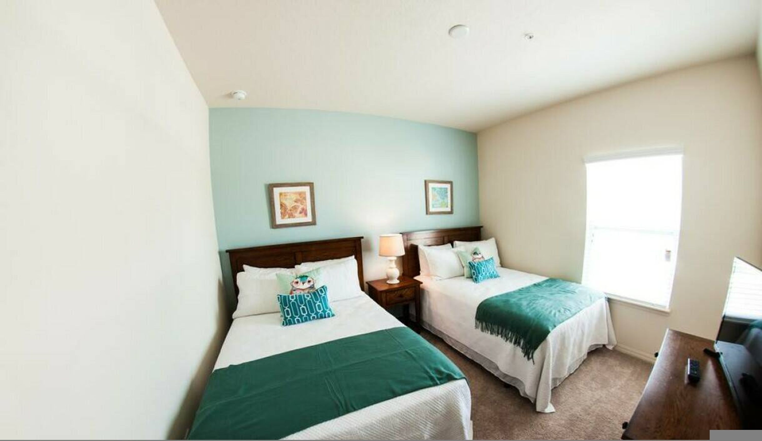 A hotel room with two beds, each with a green bedspread and matching pillows. There are two bedside tables with lamps, a window with a view, and two framed pictures on the wall.