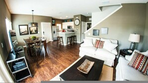 Een ruime woonkamer met een witte bank, een zwarte salontafel en een eethoek met een houten tafel en stoelen. In de achtergrond is een keuken te zien met een wit schiereiland en barstoelen.