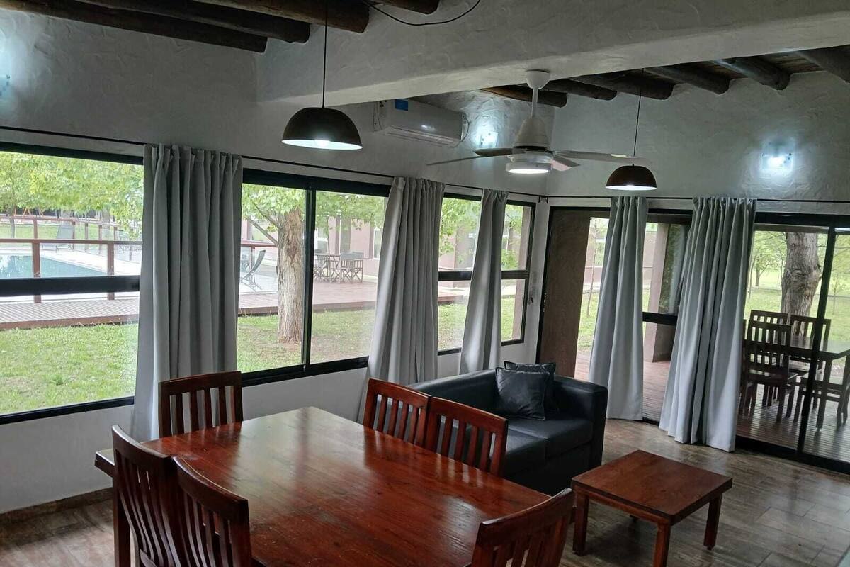 A spacious room with a wooden dining table, a sofa, and a small coffee table. Large windows with curtains allow natural light and views of the outdoors.