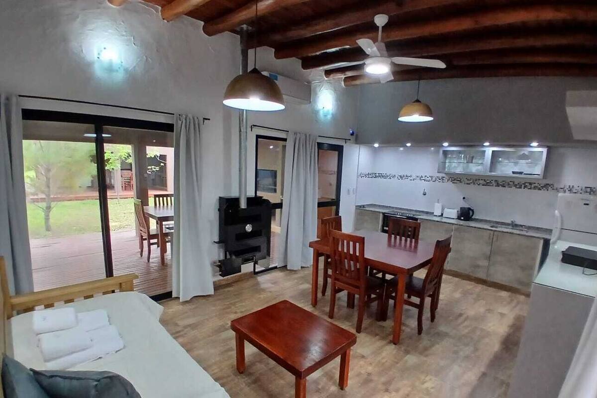 A cozy living area with a sofa, wooden coffee table, and dining table with chairs. A kitchen with a TV and a grill is visible in the background.