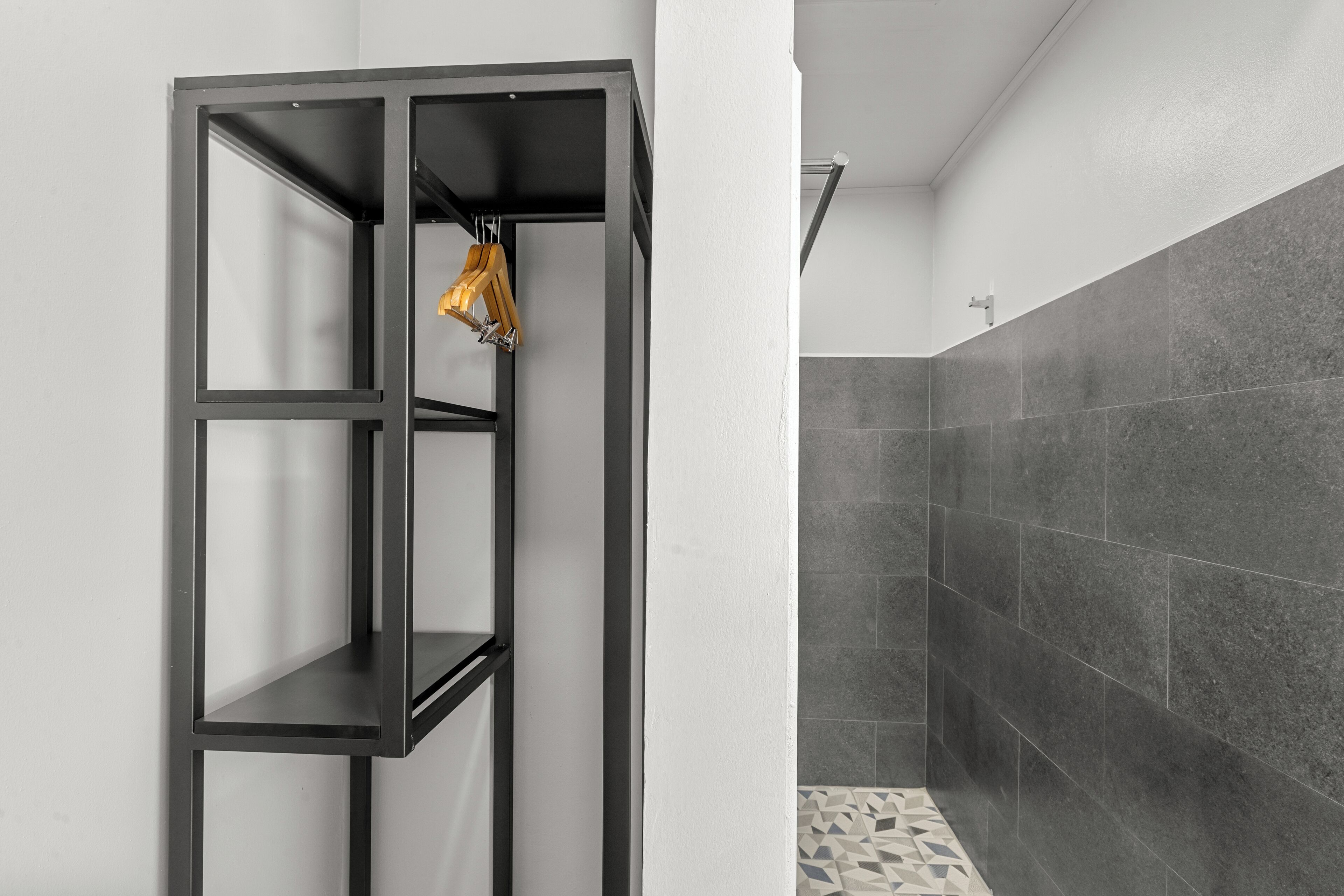 Un baño con un estante metálico negro, una ducha con baldosas oscuras y un suelo estampado.