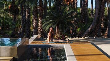 Zona de piscina con terraza de madera, palmeras y una persona meditando.