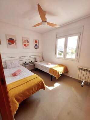 A bedroom with two beds, a ceiling fan, artwork on the wall, and a window with a view of buildings. - Stunning 2 bed Villa With Private Salt Water Pool (Torre-Pacheco)