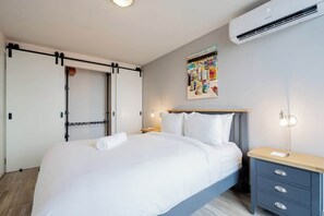 A hotel room with a bed, bedside table, wall-mounted air conditioning unit, and a wall art piece.