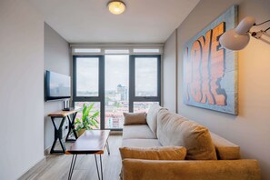 A modern living room with a sofa, a small table, a plant, and a large window offering a city view.