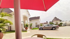 Spiseområde med et bord under en rød parasol, en bil i baggrunden og en bygning med hvid facade og stråtækt tag.