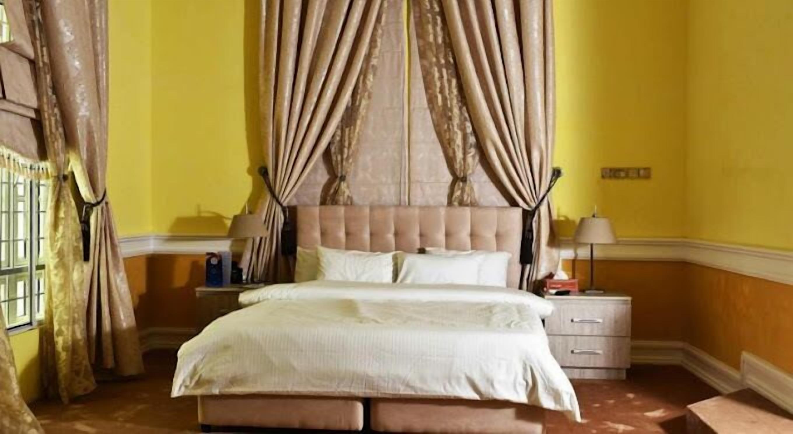 A bedroom with a tufted headboard, a bed with white linens, two bedside tables with lamps, and curtains on the windows.