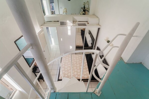 A spiral staircase leading to a bedroom with a bed, a small table, and a window.