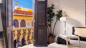 Habitación de hotel con balcón, cama con estructura blanca, mesita de noche con lámpara y vista a un edificio amarillo con tejas rojas.