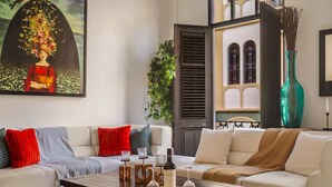 A living room with a white sofa, red and blue cushions, a coffee table with a bottle and glasses, and a painting on the wall.