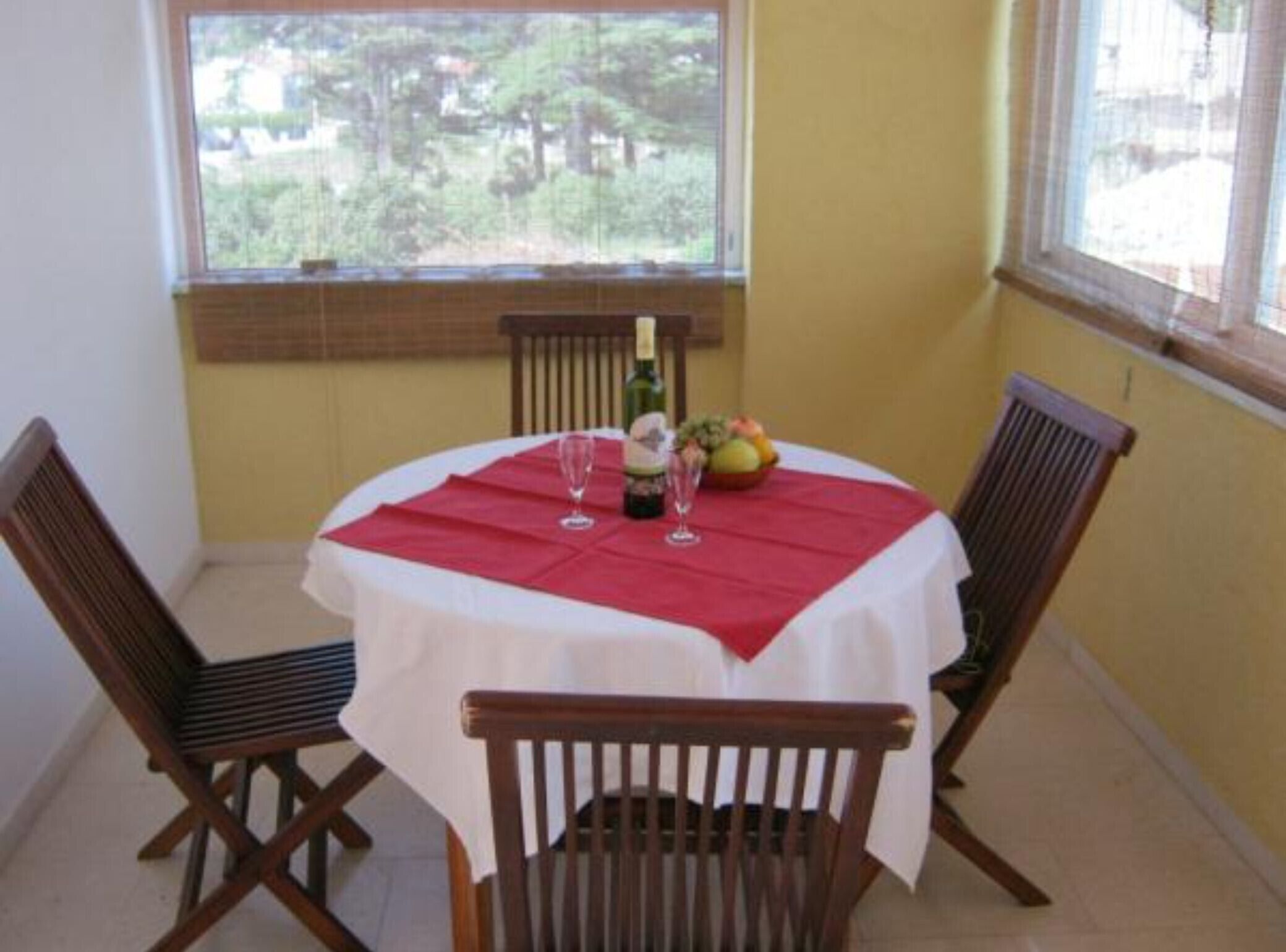 Zona pranzo con tavolo rotondo ricoperto da tovaglia bianca e runner rosso. Due sedie in legno circondano il tavolo. Sul tavolo sono posizionati una bottiglia di vino e due calici. Si vede anche una ciotola di frutta. La stanza ha una finestra con vista su alberi e un edificio.