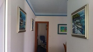 A hallway with a door leading to a room, framed artwork on the walls, and a chair in the room.