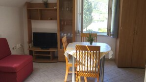 A room with a red sofa, a wooden dining table with chairs, a TV on a stand, and a window with blinds.