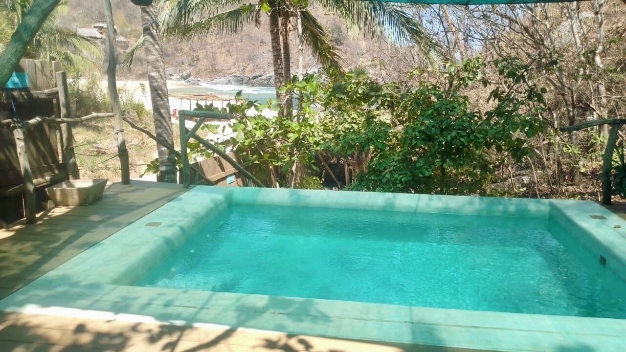 Piscina rectangular al aire libre con agua cristalina, rodeada de vegetación y vistas a una playa y edificios al fondo.