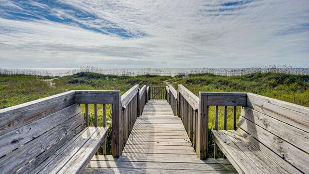 A wooden boardwalk leading to a beach with a grassy area and a cloudy sky. - Home on Ocean Isle Beach w/ Catwalk & Deck (Ocean Isle Beach)