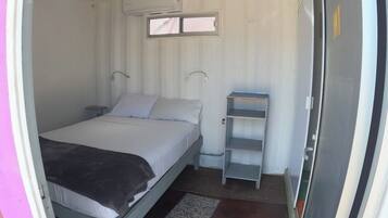 Ein kleines Zimmer mit einem Einzelbett, einem schmales Fenster, einem Regal und einer Tür.