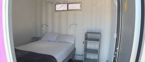 Habitación compacta con cama individual, una ventana pequeña, estante y puerta.