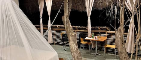 Un espacio al aire libre con carpas, una mesa y sillas para comer, rodeado de árboles y una estructura de techo de paja.