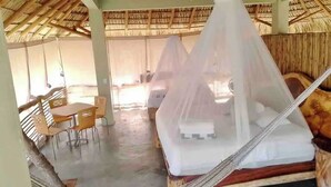 Una habitación con una cama cubierta por una mosquitera, una mesa con sillas y un techo de paja.