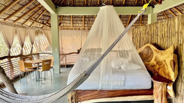 Una hamaca con mosquitero, cabecera de madera y zona de comedor con sillas y una mesa.