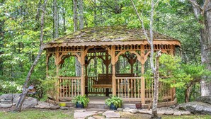 A wooden gazebo with a thatched roof, surrounded by a stone pathway and potted plants, set in a forested area. - Cozy Family Cabin in charming Smoky Mountain- Pets allowed  (Cosby)