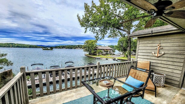 A deck with a view of a lake, a dock with boats, and a small building with an anchor decoration. - Farr Shores Cozy Retreat (Hot Springs)