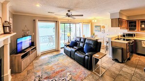 A living room with a black leather couch, a television, and a kitchen area with wooden cabinets. - Farr Shores Cozy Retreat (Hot Springs)