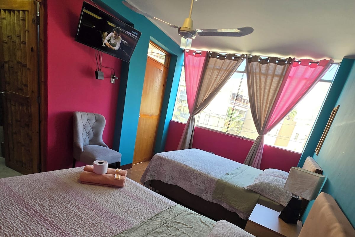 A room with two beds, a television, a ceiling fan, and a window with curtains.
