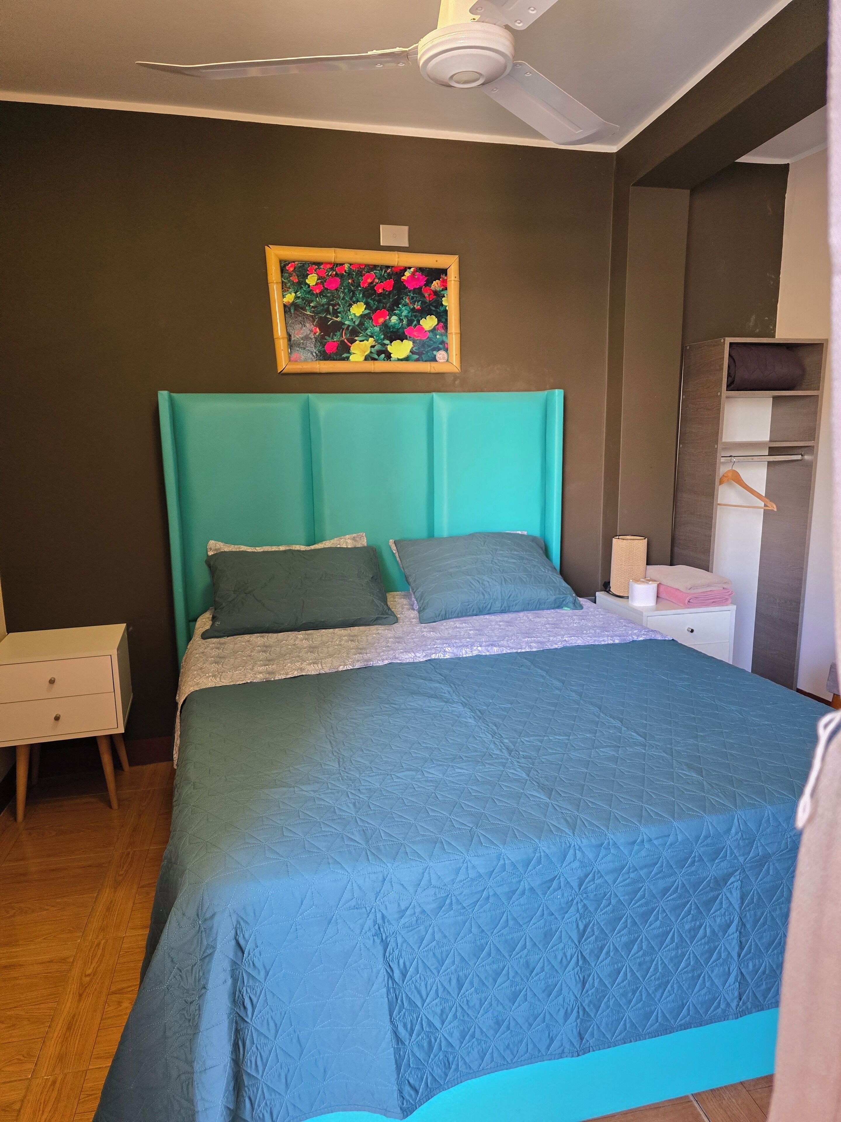 A bedroom with a teal headboard, a bed with a blue quilt, a nightstand, a bedside table, and a wall-mounted shelf with a rolled-up towel and a hanging garment.