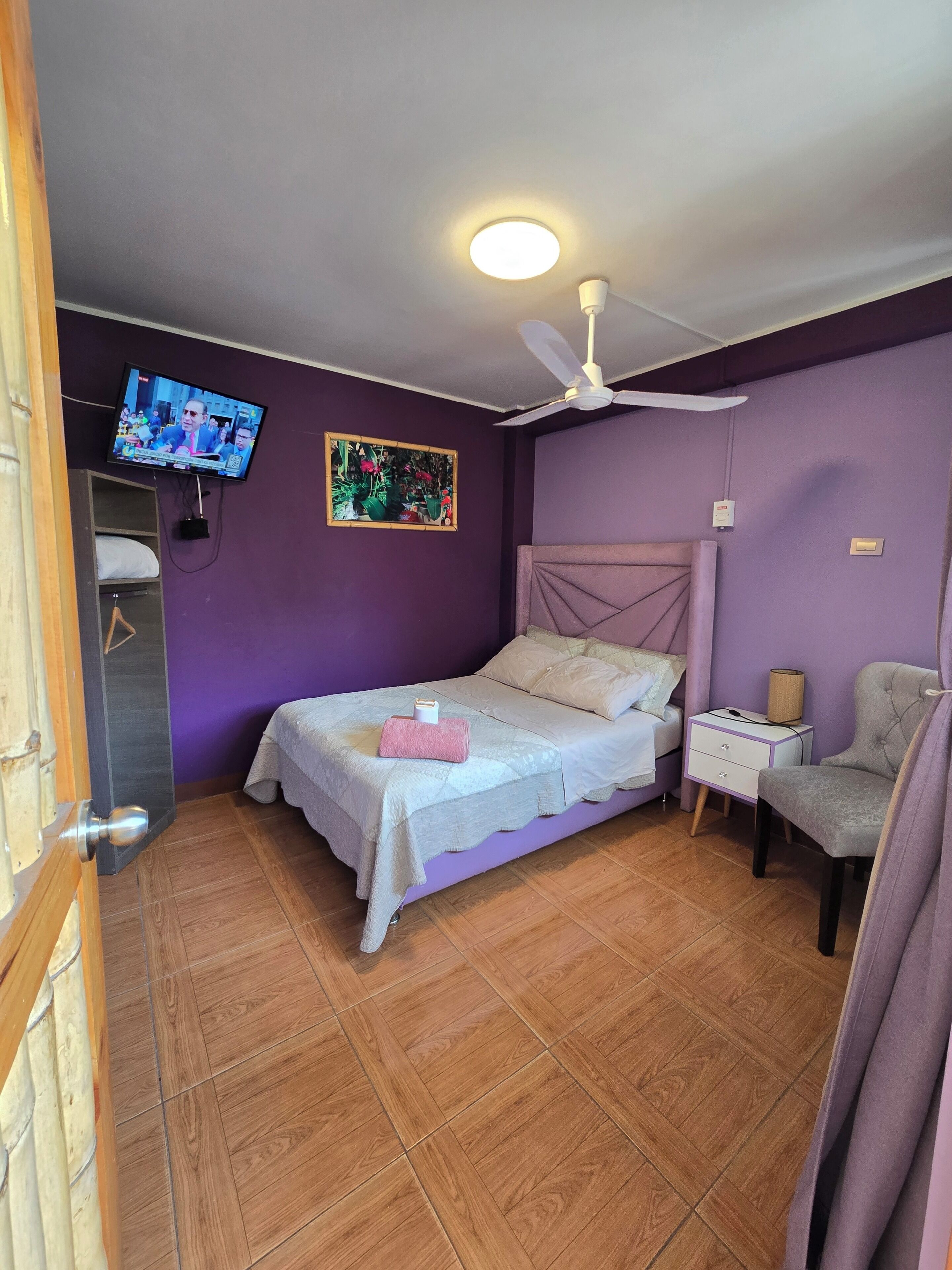 Un dormitorio con cama, mesita de noche, silla, ventilador de techo, televisor y un cuadro en la pared.