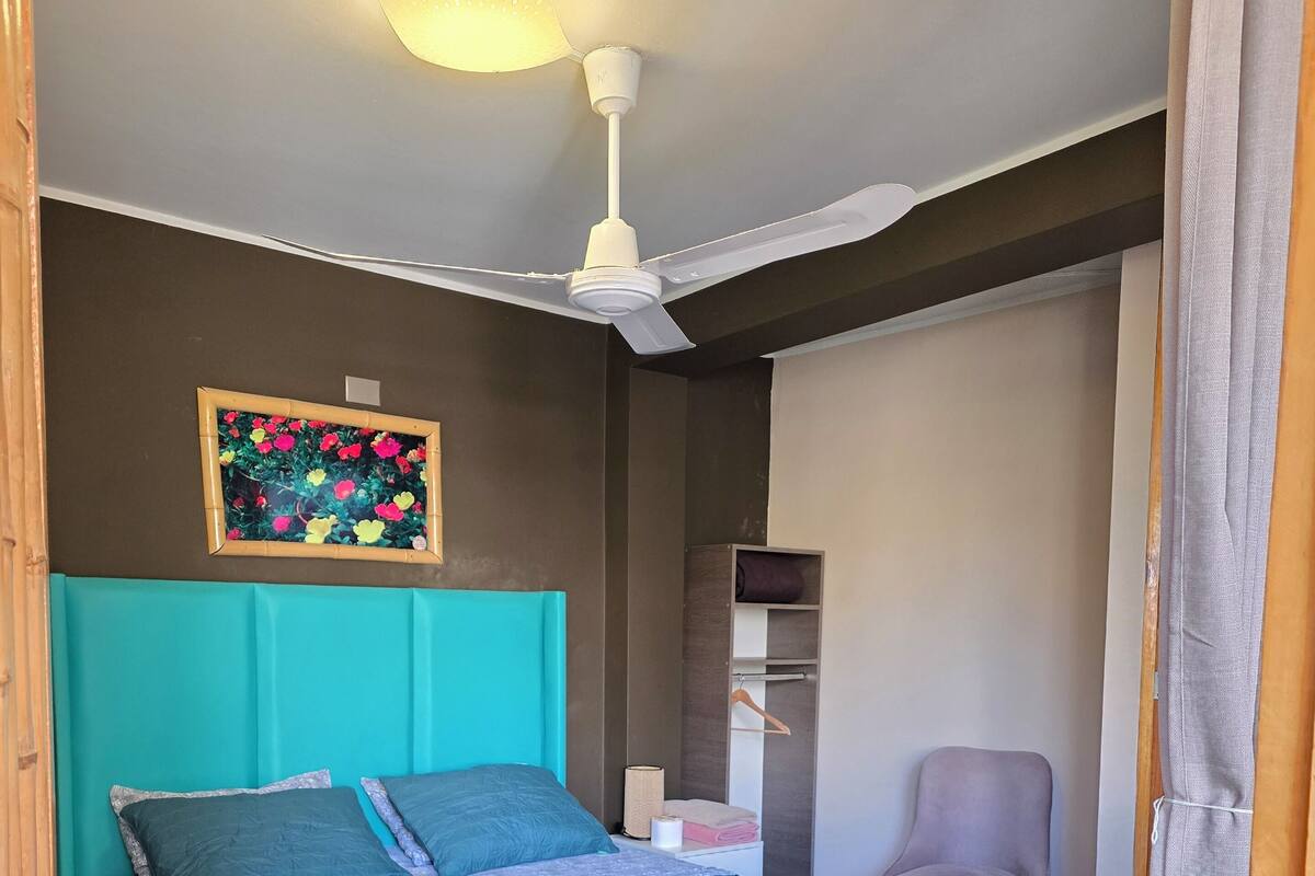 A bedroom with a bed, a ceiling fan, a wall-mounted shelf, and a chair.