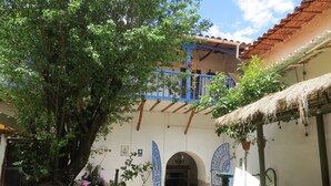 Un patio con un muro de piedra, un árbol grande, una puerta azul y una estructura de techo de paja.