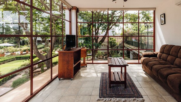 Una habitación amplia con ventanas grandes, un ventilador de techo, un mueble de televisión de madera, un sofá marrón y una mesita pequeña.