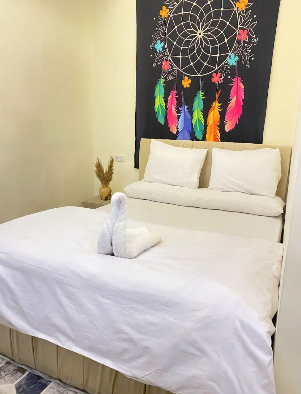 A neatly made bed with white bedding and a decorative towel, a wall-mounted dreamcatcher with colorful feathers, and a small vase with dried plants on a bedside table.