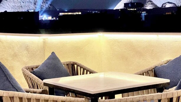 Área de estar externa com cadeiras de vime e uma mesa, com vista para as pirâmides à noite.