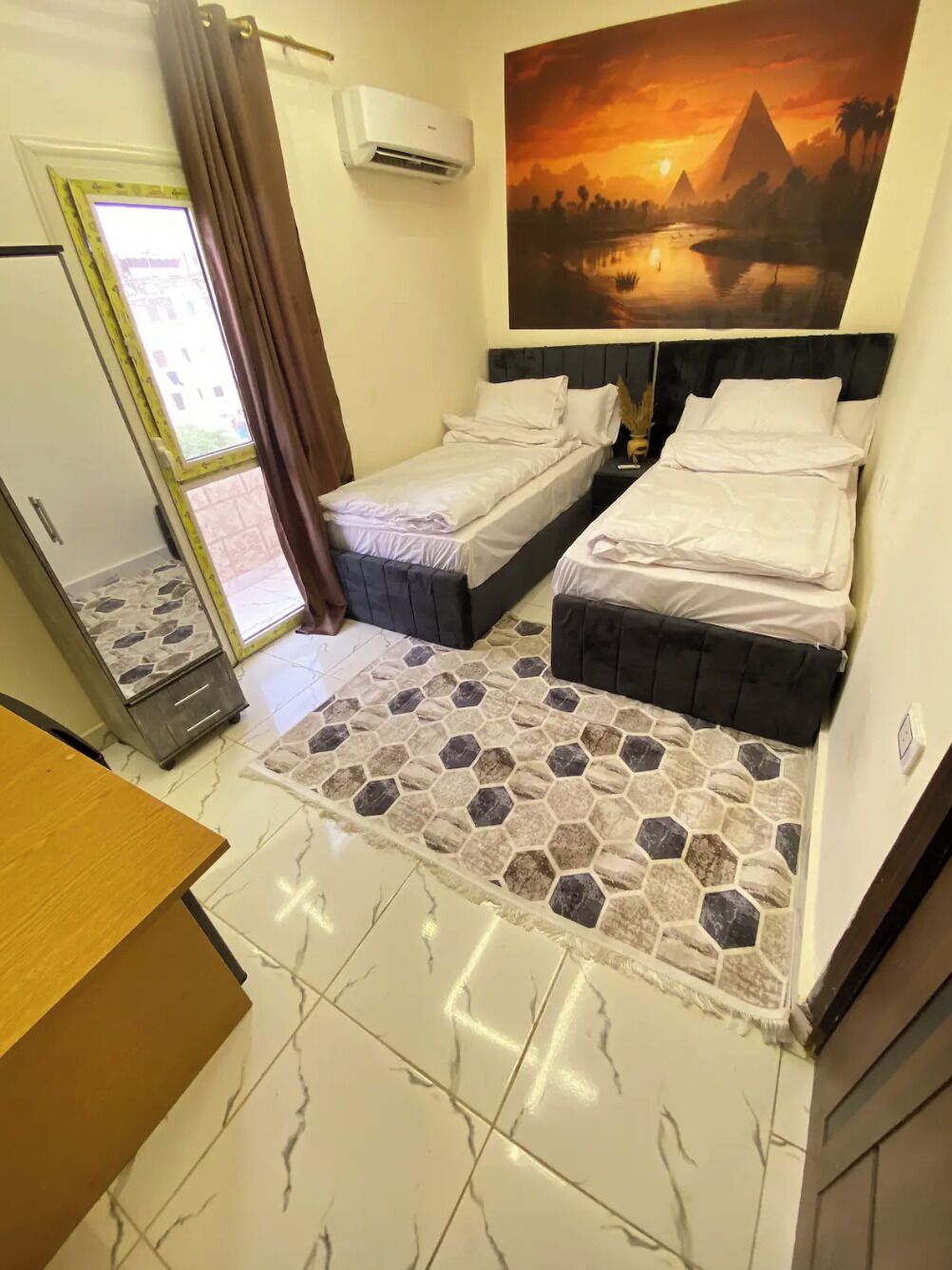 A bedroom with two beds, a stone-patterned rug, a large painting of a sunset, and a window with curtains.